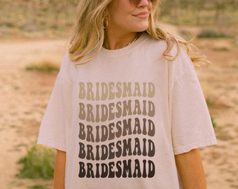 May include: Cream-colored t-shirt with the word "BRIDESMAID" stacked five times in a wavy, gradient design. The text transitions from light beige to dark brown. The shirt is worn outdoors with a desert landscape in the background.