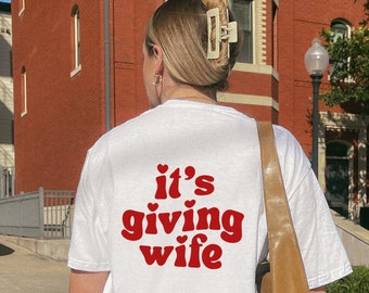 May include: White t-shirt with the text "it's giving wife" in red, heart-shaped font. The shirt is worn by a person with blonde hair, a beige hair clip, and gold hoop earrings. A brown shoulder bag is also visible.