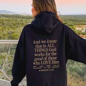 May include: Black hoodie with gold text on the back. The text reads "And we know that in ALL THINGS God works for the good of those who LOVE Him", with a reference to Romans 8:28. The hoodie has a hood and long sleeves.