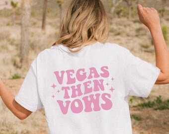 May include: White t-shirt with pink text that reads "Vegas Then Vows" in a retro font. The shirt is being worn by a person in a desert landscape.
