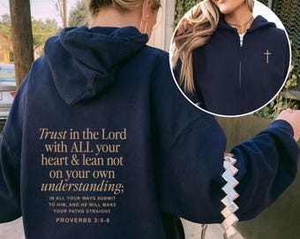 May include: Navy blue hoodie with the text "Trust in the Lord with ALL your heart & lean not on your own understanding;" and "Proverbs 3:5-6" printed on the back. A small gold cross is on the front. The hoodie is worn with blue jeans.