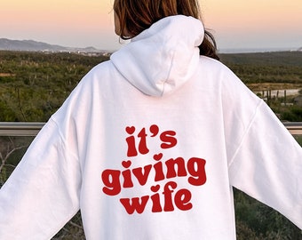 May include: White hoodie with red text that says "it's giving wife" with small hearts around the text.