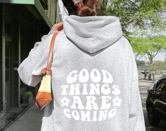 May include: A gray hooded sweatshirt with the text "GOOD THINGS ARE COMING" in white lettering with a flower design. The sweatshirt is being worn by a person walking down a city street.