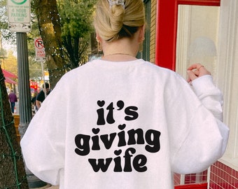 May include: White sweatshirt with black text that reads "it's giving wife" with small hearts around the text.
