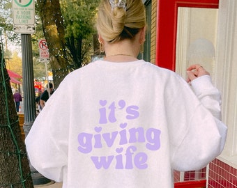 May include: White sweatshirt with a purple graphic that says "it's giving wife" with hearts around the text.