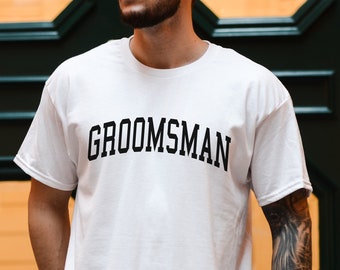 May include: White t-shirt with the word "GROOMSMAN" printed in black letters.