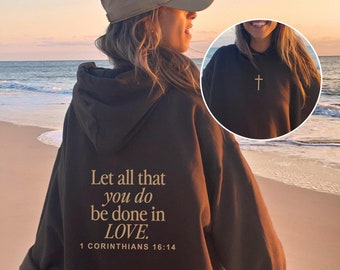 May include: A brown hooded sweatshirt with the text "Let all that you do be done in LOVE. 1 CORINTHIANS 16:14" printed on the back. The model is wearing the sweatshirt with a beige baseball cap and is standing on a sandy beach.