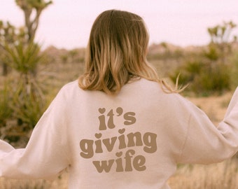May include: A beige sweatshirt with the phrase "it's giving wife" printed on the back in brown lettering. The sweatshirt is worn by a person standing in a desert landscape with Joshua trees and a pinkish sky.