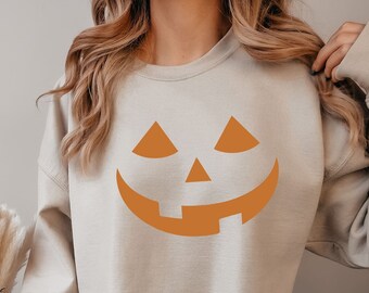 May include: Beige sweatshirt featuring a jack-o'-lantern face in orange. The pumpkin face has triangular eyes, a triangular nose, and a wide, smiling mouth. The sweatshirt is a neutral color, suitable for fall and Halloween.