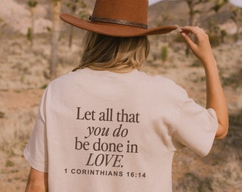 May include: A person wearing a beige t-shirt with the text "Let all that you do be done in LOVE. 1 Corinthians 16:14" printed on the back. The person is wearing a brown cowboy hat and is standing in a desert landscape.