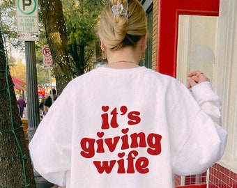 May include: White sweatshirt with red text that says "it's giving wife" with a heart symbol above the word "it's" and below the word "giving".
