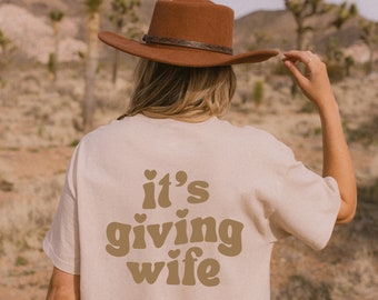 May include: A beige t-shirt with the text "it's giving wife" in brown letters with hearts above the "it's" and below the "wife".