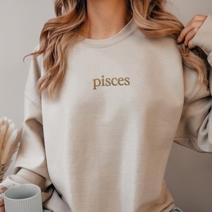 May include: A light beige sweatshirt with the word "pisces" embroidered in gold on the front.