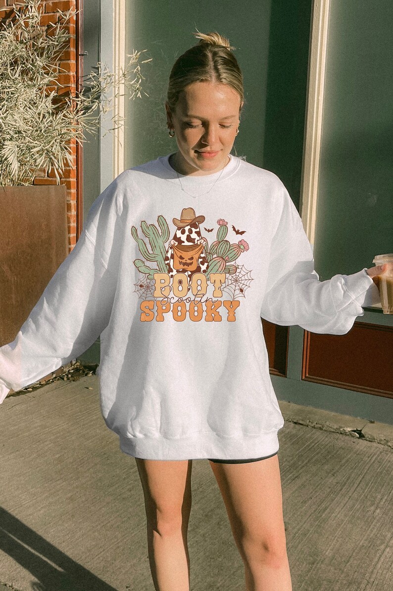May include: White sweatshirt with a graphic of a cowboy cactus holding a pumpkin bucket with the text "Boot Scootin' Spooky".