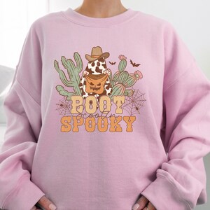May include: Pink sweatshirt featuring a graphic of a ghost wearing a cowboy hat and holding a pumpkin bucket. The ghost is surrounded by cacti and bats. The text "Boot Scootin' Spooky" is printed below the ghost.