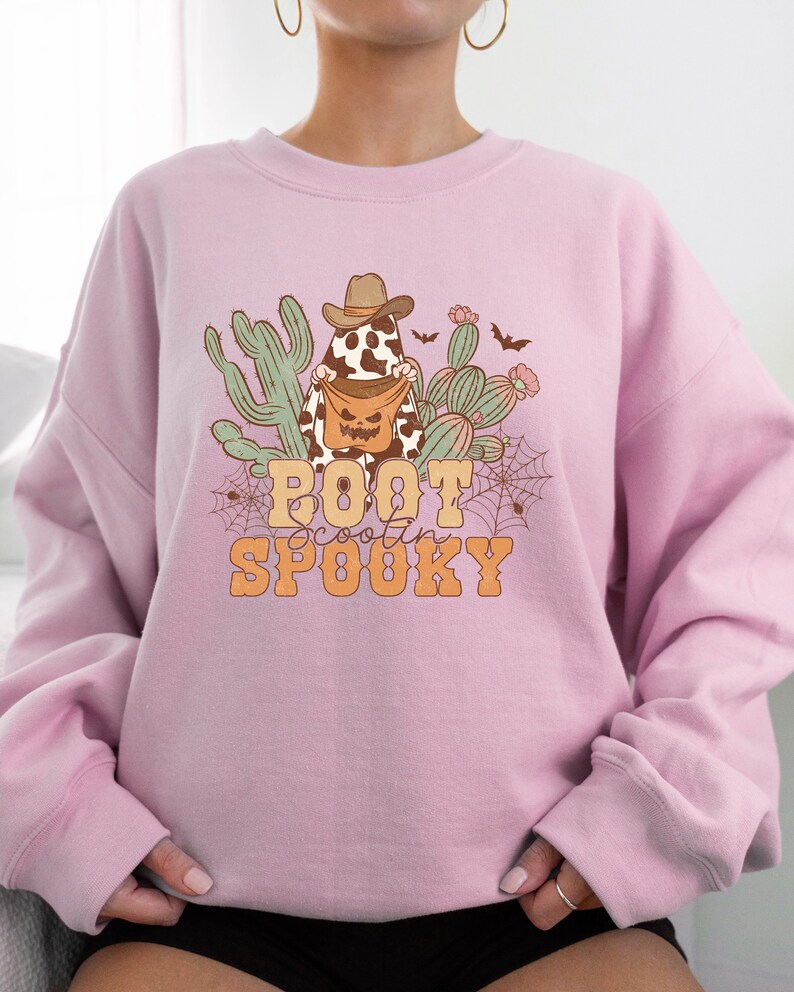 May include: Pink sweatshirt featuring a graphic of a ghost wearing a cowboy hat and holding a pumpkin bucket. The ghost is surrounded by cacti and bats. The text "Boot Scootin' Spooky" is printed below the ghost.