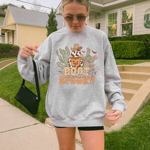May include: Grey sweatshirt with a graphic of a cartoon cowboy boot holding a jack-o-lantern with the text "Boot Scootin' Spooky".