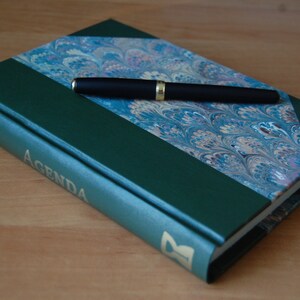 May include: A green leather-bound journal with a marbled paper design on the front cover. The journal has the word "AGENDA" printed in gold lettering on the spine. A black pen is lying on top of the journal.