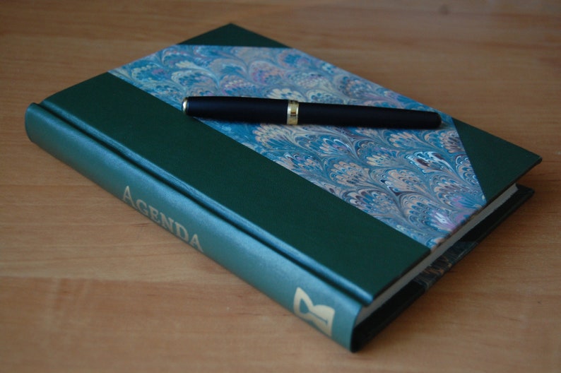 May include: A green leather-bound journal with a marbled paper design on the front cover. The journal has the word "AGENDA" printed in gold lettering on the spine. A black pen is lying on top of the journal.