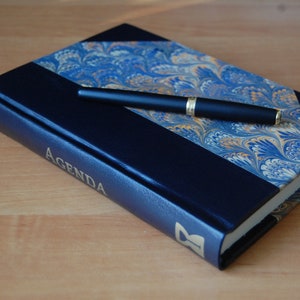May include: A blue leather-bound journal with the word "AGENDA" embossed in gold on the cover. A black pen is lying on top of the journal.