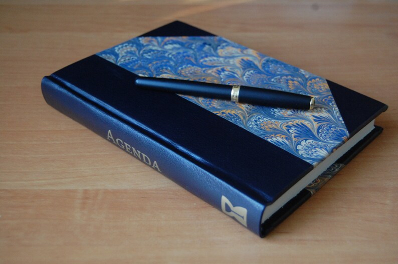 May include: A blue leather-bound journal with the word "AGENDA" embossed in gold on the cover. A black pen is lying on top of the journal.