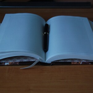 May include: An open lined notebook with a black pen lying on the pages. The notebook has a brown cover with a decorative pattern.