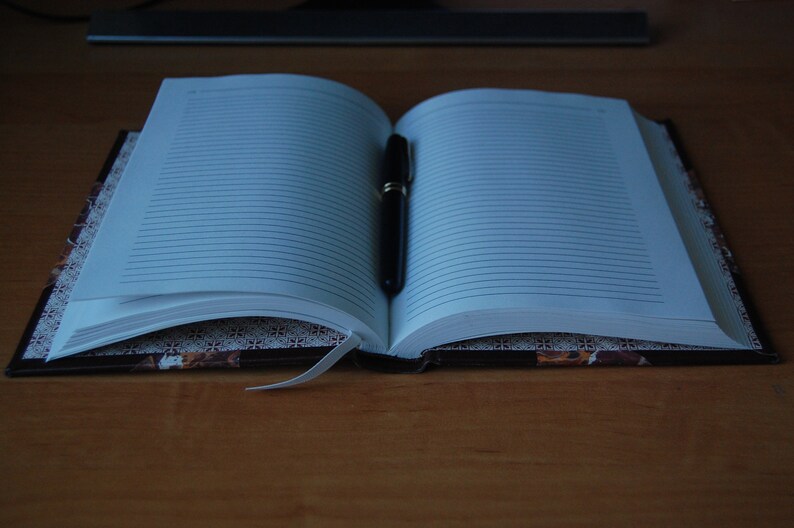 May include: An open lined notebook with a black pen lying on the pages. The notebook has a brown cover with a decorative pattern.