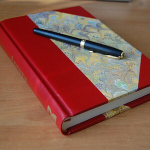 May include: A red leather-bound journal with a marbled paper design on the front cover. A black pen is lying on top of the journal.