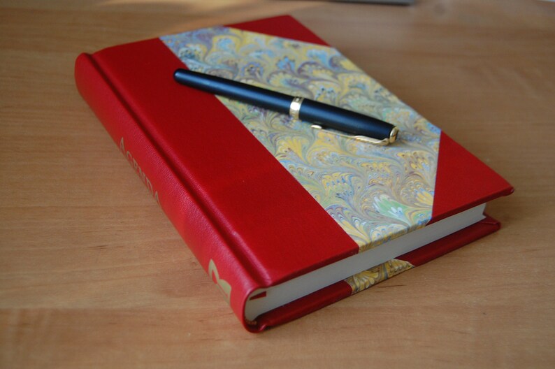 May include: A red leather-bound journal with a marbled paper design on the front cover. A black pen is lying on top of the journal.