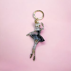 May include: A gold key chain with a black and white ballerina charm. The ballerina is in a mid-air pose with her arms outstretched.