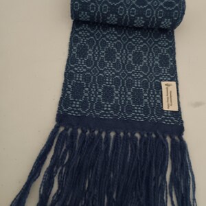 May include: A rolled-up, dark blue scarf with a woven floral pattern and fringe. The scarf is made of a soft material and features a tag that reads "Handmade by Spinning Sheep Crafts".