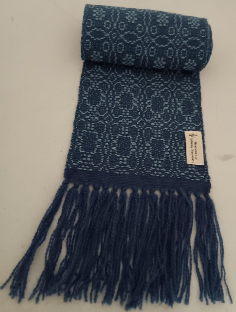 May include: A rolled-up, dark blue scarf with a woven floral pattern and fringe. The scarf is made of a soft material and features a tag that reads "Handmade by Spinning Sheep Crafts".