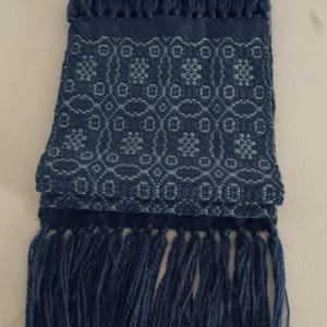 May include: A folded, indigo blue scarf with a geometric pattern and fringe. The scarf features a repeating floral design in a lighter shade of blue. The ends of the scarf are finished with long, thick fringe.