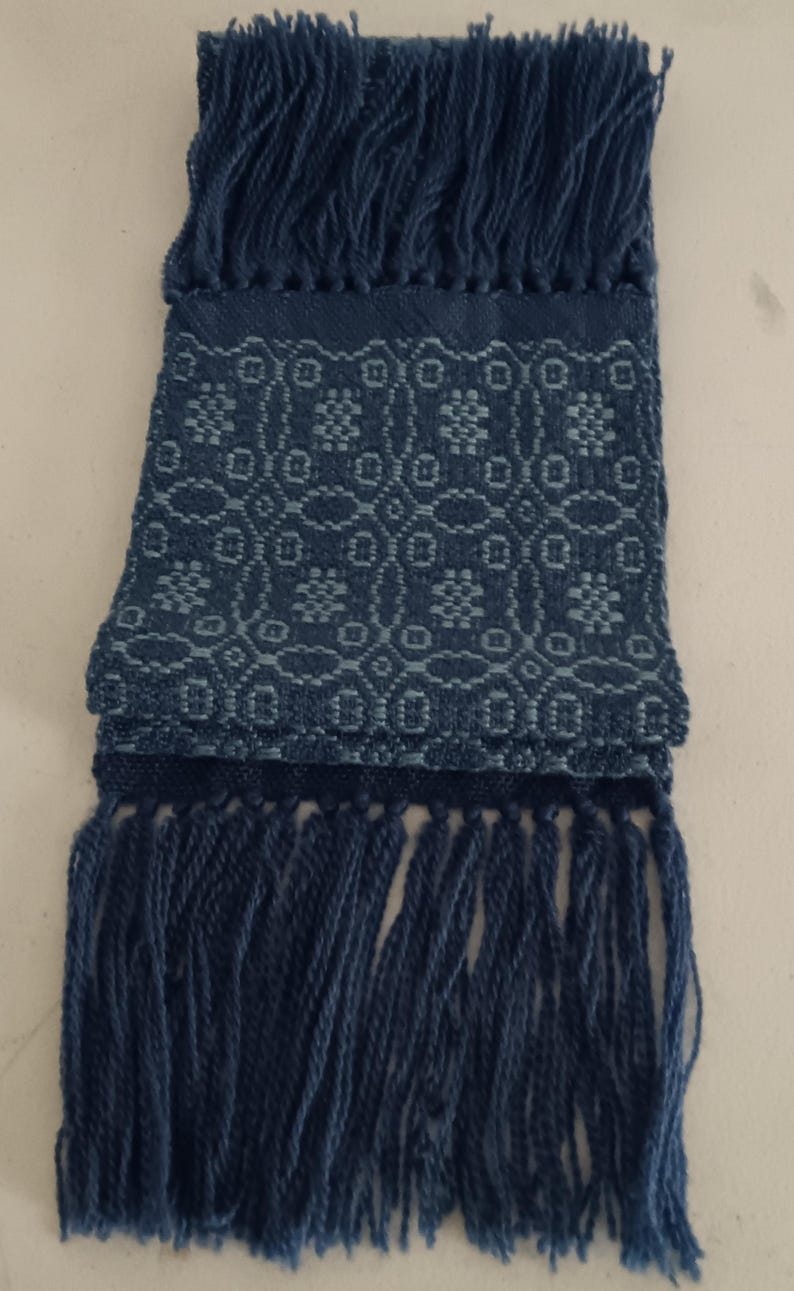 May include: A folded, indigo blue scarf with a geometric pattern and fringe. The scarf features a repeating floral design in a lighter shade of blue. The ends of the scarf are finished with long, thick fringe.