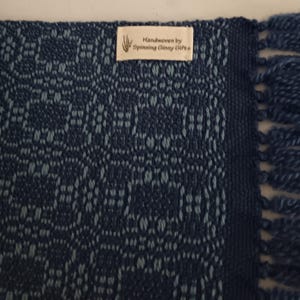 May include: A handwoven textile in shades of blue, featuring a geometric pattern. The edge has a dark blue border and fringe. A tag reads "Handwoven by Spinning Ginny Gifts."