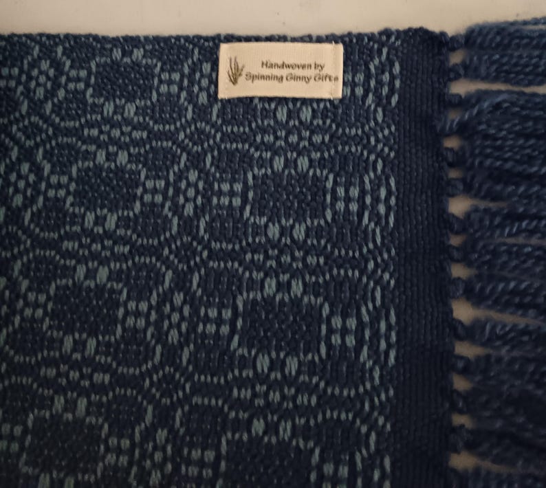 May include: A handwoven textile in shades of blue, featuring a geometric pattern. The edge has a dark blue border and fringe. A tag reads "Handwoven by Spinning Ginny Gifts."