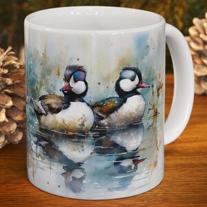 Könnte beinhalten: Weiße Keramiktasse mit einem Aquarellgemälde von zwei Mandarinenten auf einem See. Die Enten haben ein farbenfrohes Gefieder mit weißen, schwarzen und braunen Federn. Die Tasse hat einen weißen Henkel und ist für Heißgetränke geeignet.