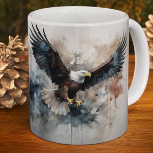 May include: White ceramic mug with a watercolor illustration of a bald eagle in flight against a cloudy background. The eagle's wings are spread wide, and its talons are extended. The background is a soft gray with splashes of color.