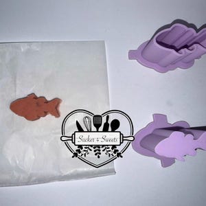 May include: A baked fish-shaped cookie on parchment paper next to two purple fish-shaped cookie cutters. The cookie cutters are made of plastic and are different sizes. The image also includes a logo that says "Sucker + Sweets".
