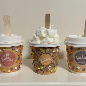May include: Three decorated paper cups with white lids and wooden stirrers. The cups are brown with colorful sprinkles and circular labels with the text "Sucker" and "Sweets". One cup is filled with marshmallows.