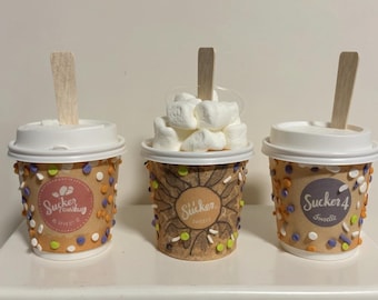 May include: Three decorated paper cups with white lids and wooden stirrers. The cups are brown with colorful sprinkles and circular labels with the text "Sucker" and "Sweets". One cup is filled with marshmallows.
