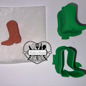 May include: An image features a baked cookie shaped like a cowboy boot, alongside two green plastic boot-shaped cookie cutters. The cookie is on parchment paper. The logo "Sucker 4 Sweets" is also visible.