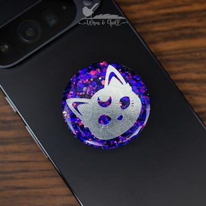 Peut inclure: Un support de téléphone rond avec un motif de visage de chat argenté sur un fond de paillettes violettes. L'accessoire de téléphone est fixé à un smartphone noir. La marque "Wren & Quill" est visible en haut à gauche.