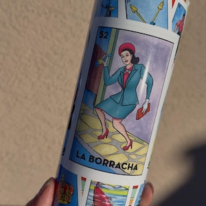 Può includere: Un bicchiere bianco con un design colorato che presenta una donna in un abito blu e il testo "La Borracha" su uno sfondo blu. Il design è ispirato alle tradizionali carte messicane di Loteria.