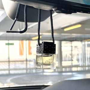 May include: A car air freshener with a clear glass bottle and a black, decorative top. The bottle contains a light yellow liquid and hangs from a black cord. The text "Car Secret" is visible on the bottle.