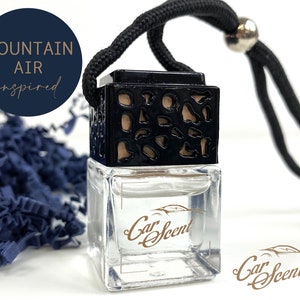 May include: A black and clear glass car air freshener with a black string hanging from it. The air freshener has a decorative wooden top and a label that says "Car Scent". The air freshener is on a white background with blue paper shreds. The text "Mountain Air inspired" is in a circle above the air freshener.