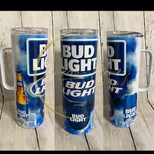 Pode incluir: Tr&ecirc;s canecas de a&ccedil;o inoxid&aacute;vel com tema Bud Light com al&ccedil;as brancas. As canecas apresentam o logotipo Bud Light em branco sobre um fundo azul com um design de nuvens. Uma caneca tem um gr&aacute;fico de garrafa Bud Light.