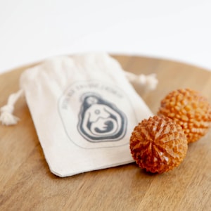 May include: Two round, textured brown wooden massage balls with a carved geometric pattern, accompanied by a small off-white cotton drawstring pouch featuring a dark grey graphic illustration of a stylized figure holding a circular object.