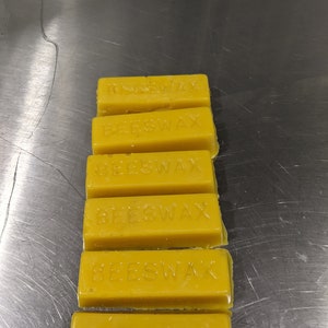 May include: Six yellow beeswax blocks stacked on top of each other on a silver surface. Each block has the word "BEESWAX" embossed on the top.