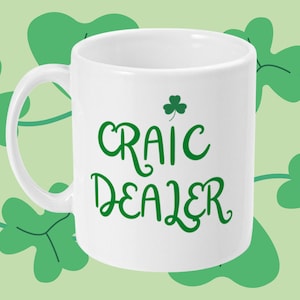 May include: White ceramic mug featuring the words "Craic Dealer" in green script, and a green shamrock. The mug has a curved handle. The background is light green with darker green shamrock shapes.